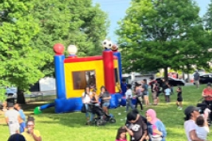 A festive outdoor event with a bounce house as the main attraction, surrounded by a diverse group of people enjoying a sunny day in the park.