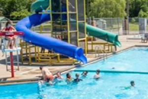 A sunny day at an outdoor public pool. A lifeguard sits on a tall chair on the left, observing children playing in the pool. Two water slides, one blue and one green, are in the background.