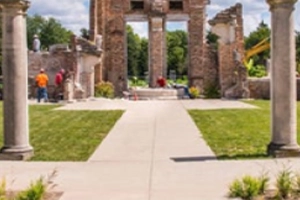 This image shows the "Ruins" at Holliday Park in Indianapolis. Workers are visible in the background, likely maintaining the grounds. The scene is captured from a ground-level perspective, looking down a paved pathway towards the ruined facade, which is framed by two large columns in the foreground. The surrounding environment includes green grass and trees, suggesting a well-maintained park setting.