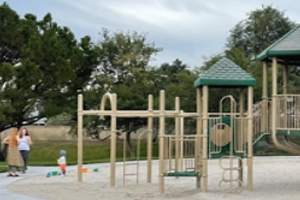 Children's playground with tan monkey bars and green slides under a cloudy sky, with two adults and a child visible in the background.